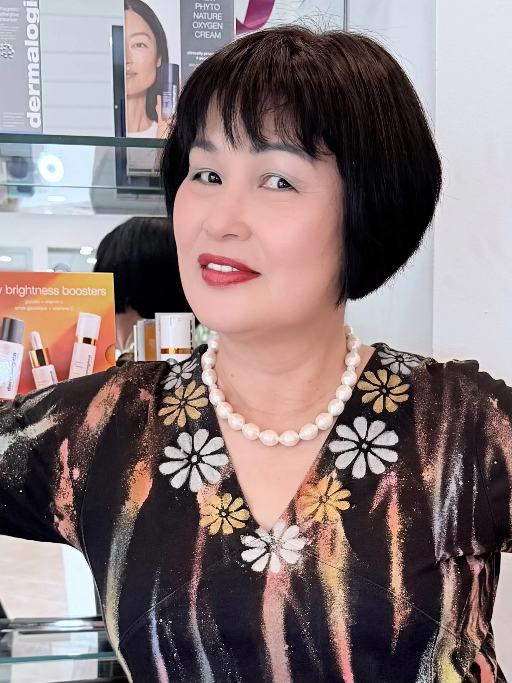 A woman with short black hair wearing a floral dress and pearl necklace.